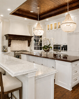 White kitchen with a marble island, crystal chandeliers, and a wood-paneled ceiling