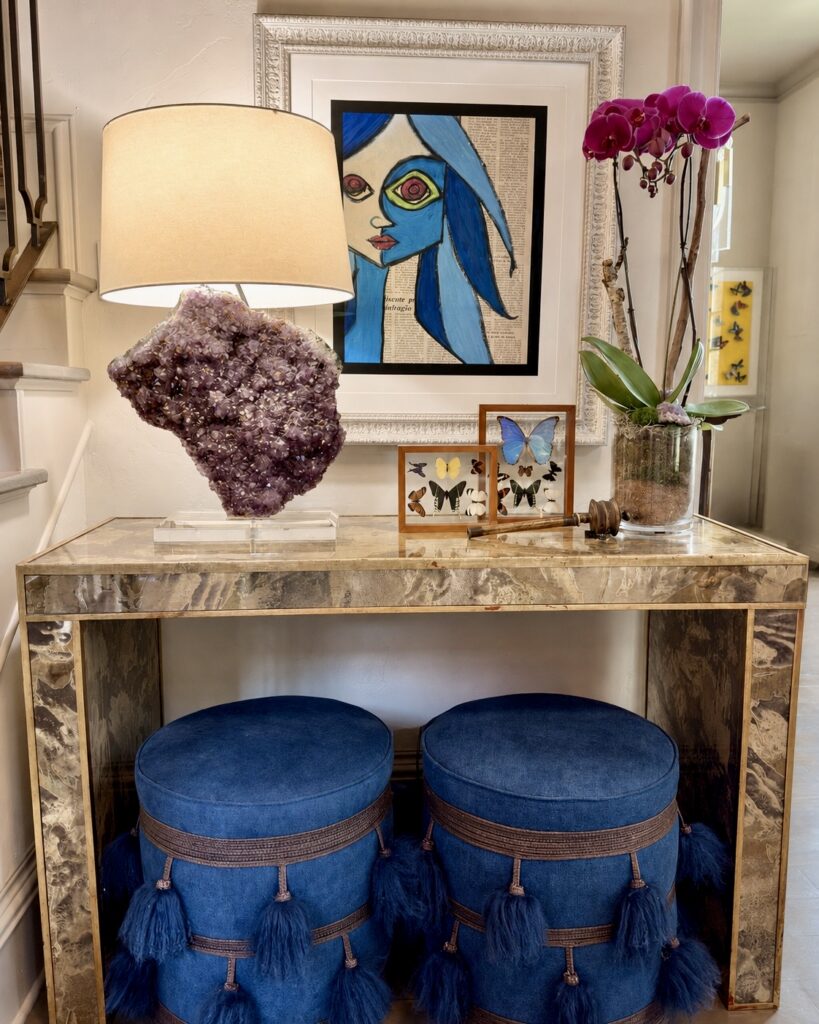 Marble console with blue tasseled ottomans, an amethyst lamp, and modern portrait art